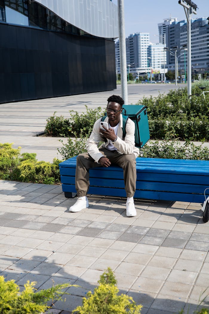 A delivery man sits on a bench outdoors, checking his phone with a thermal bag beside him.